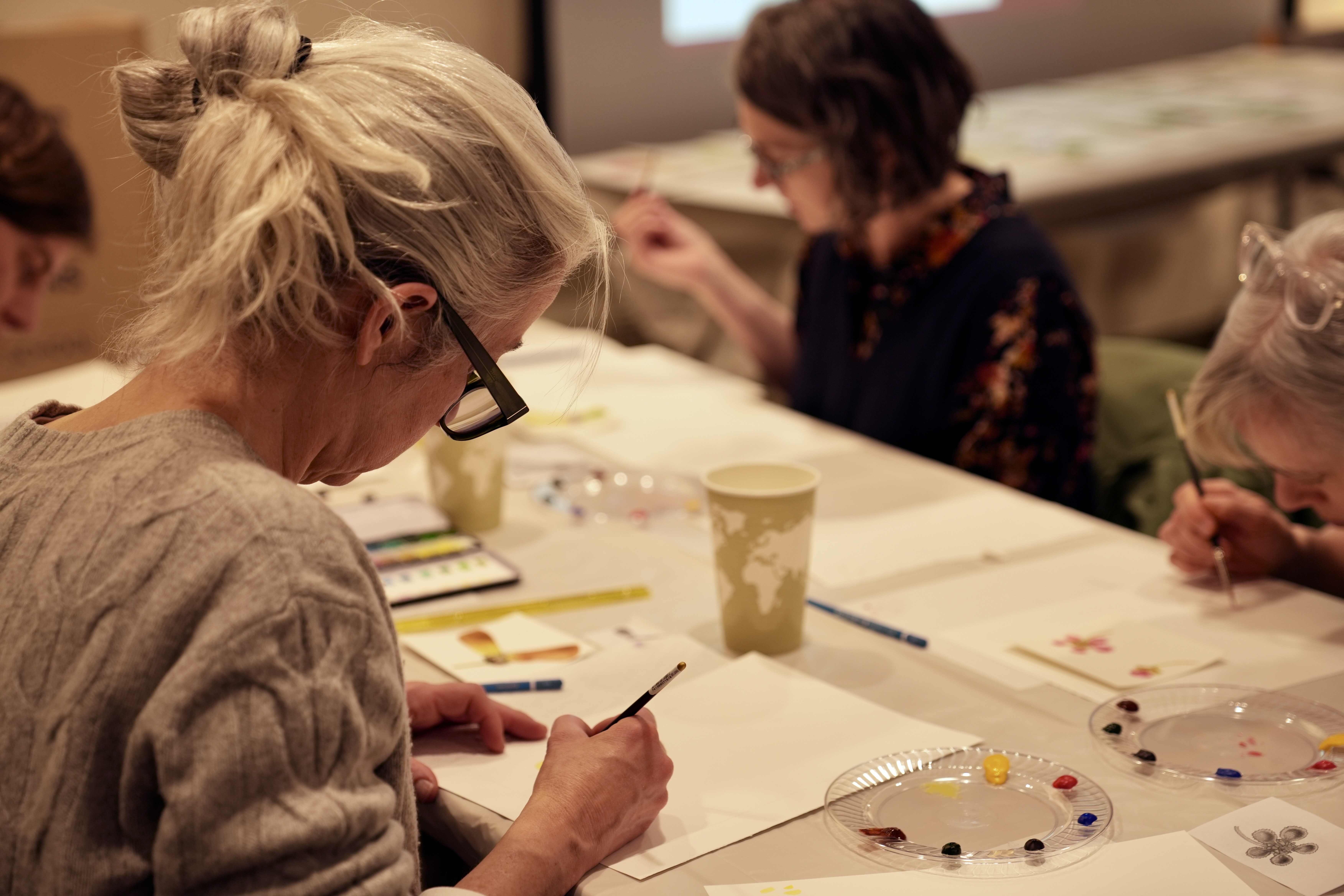 person with gray hair paints at table with other workshop participants