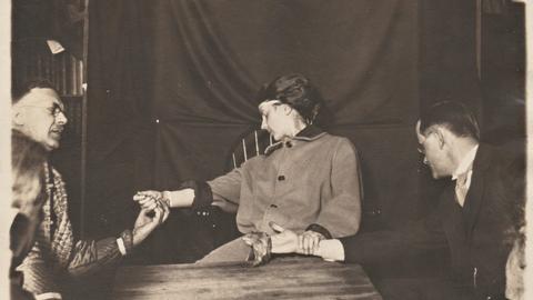 Three people participating in a seance. One woman who appears to be unconscious, men to her left and right with eyes closed. All holding hands.
