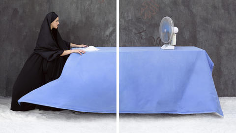 Woman in black headscarf and black robes sitting at a table in front of a pile of salt or white substance with blue tablecoth looking at the other end of the table where there is a table fan facing her.