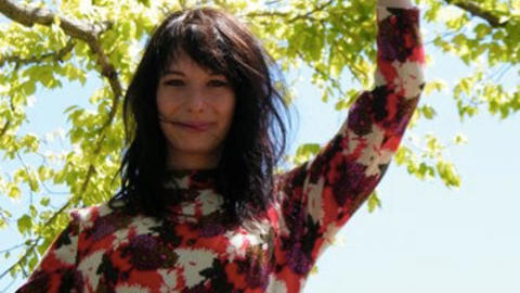 Sherah Bloor standing under a tree with arm raised, gazing at the camera