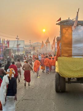 People in saris and robes walking along a busy street. Sunset in the background.