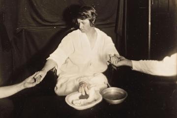 Woman seated looking down with hands holding hers on either side, a hand protuding from under her skirt, resting on table in front of her.