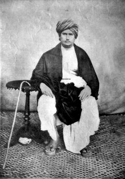 Swami Dayananda Saraswati seated next to a small table with a cane leaning on it.
