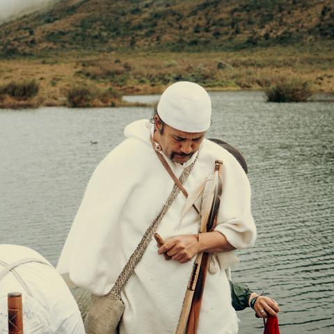 Spiritual leader clad in white standing in front of a body of water, head bowed.