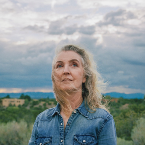Rebecca Solnit gazing skyward, brush and mountains and a clouded sky in the background