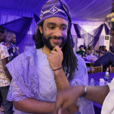 Ayodeji Ogunnaike in traditional African dress, and head piece, looking off to the side, smiling, chin in hand