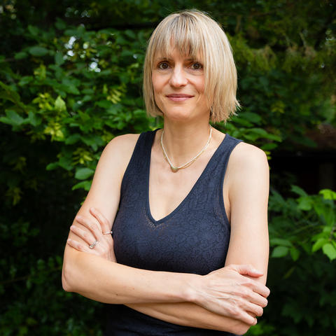 Ulrike Wagner, arms crossed standing outside in front of trees, smiling into the camera
