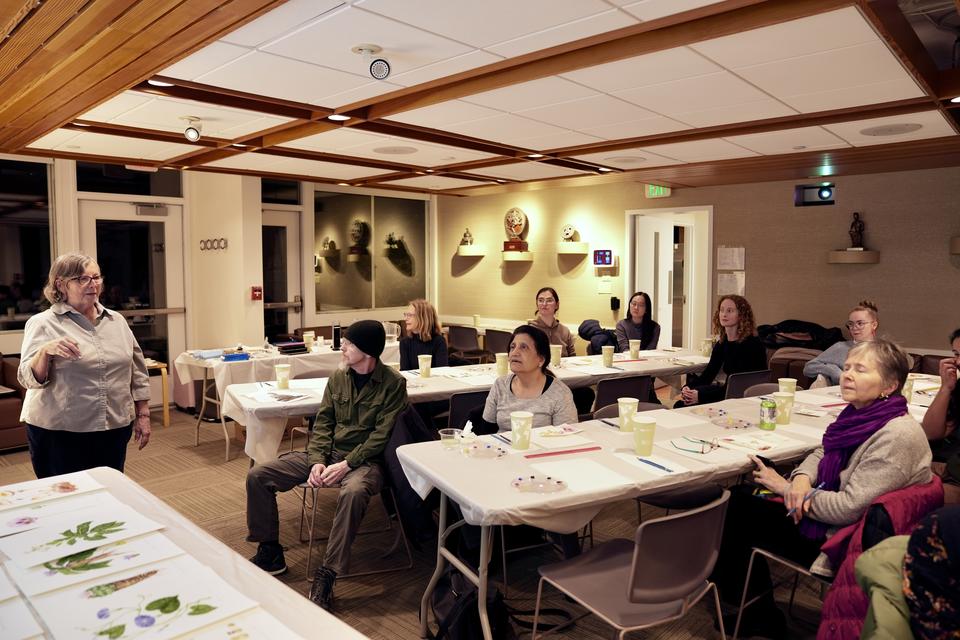 woman stands and addresses people seated at tables with painting supplies