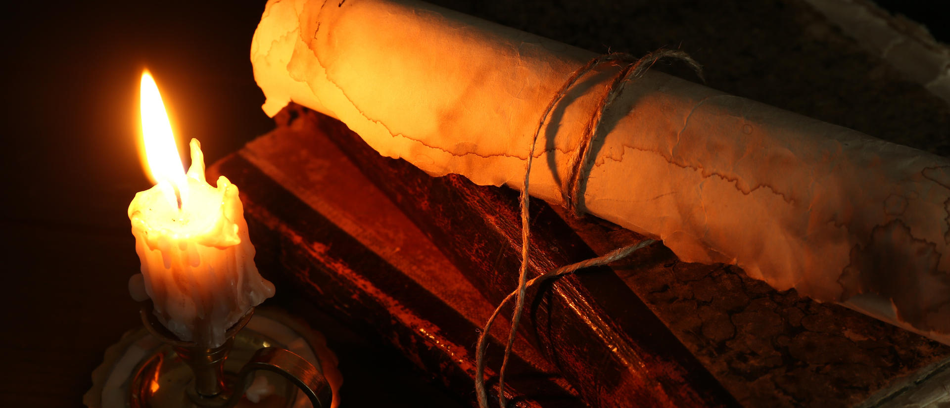 Old rolled scroll, burning candle and books on black wooden table, closeup