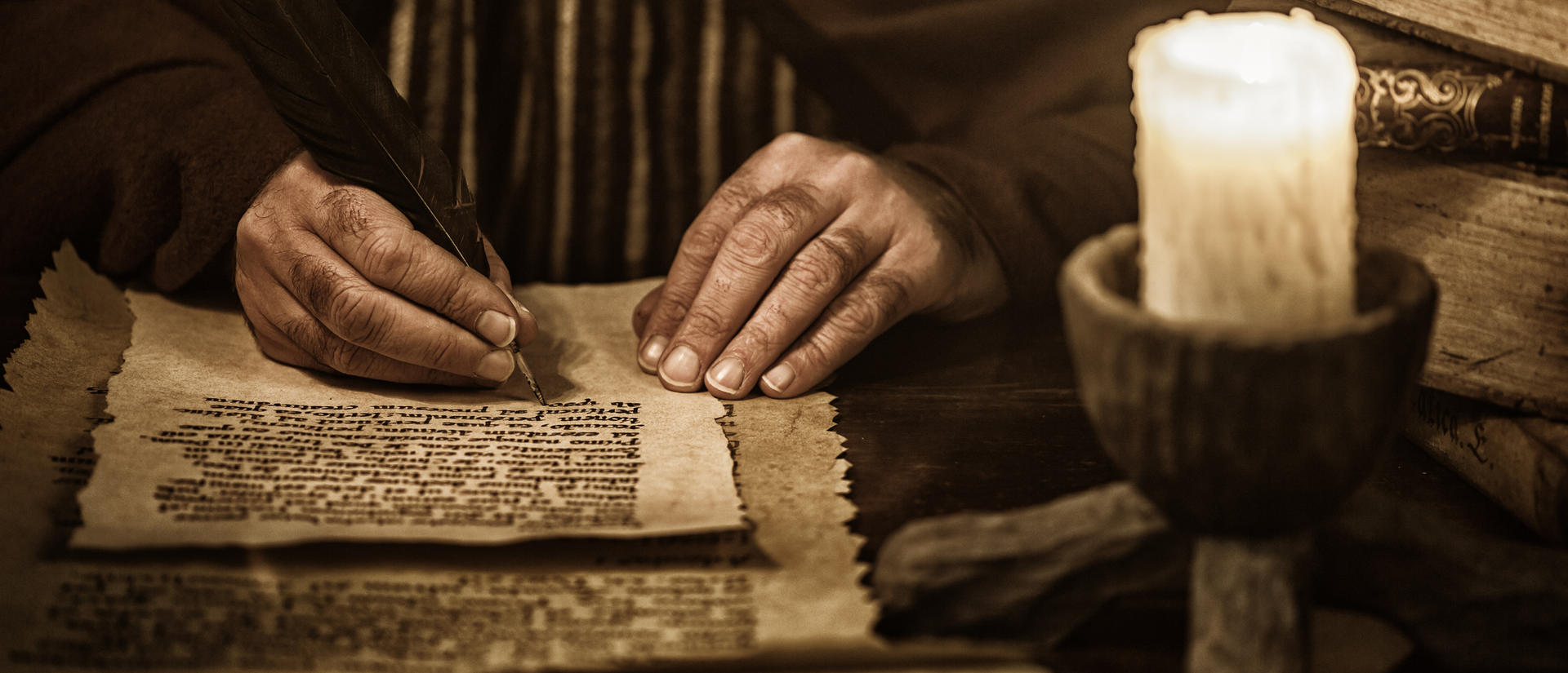 Hands writing with quill on parchment, candle close by